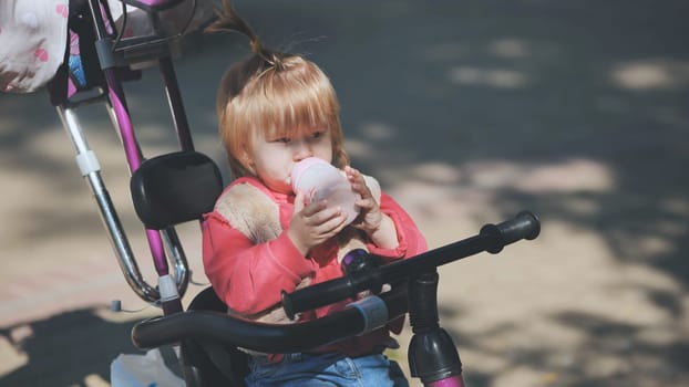 Adorable toddler girl drinking from baby bottle while sitting on her tricycle in a park, enjoying a refreshing drink on a sunny day