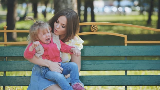 Young mother calming her crying baby daughter sitting on a bench in a park during a sunny summer day