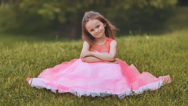 Little girl wearing beautiful pink princess dress sitting on grass in park, enjoying summer day