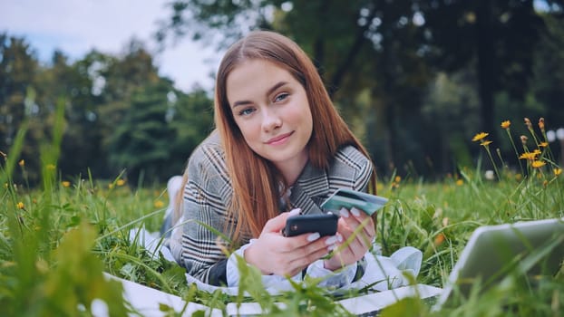 Young woman lying on grass using smartphone and credit card for online shopping in a park