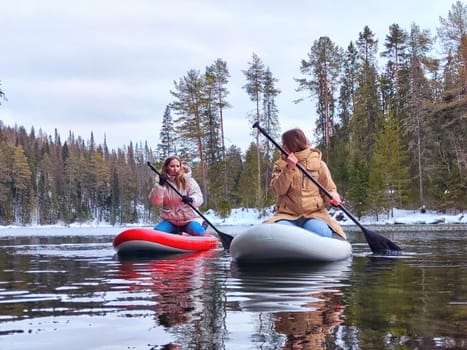 Kirov, Russia - March 14, 2024: Two women paddleboard across a serene lake, reflecting tall pine trees and winter's beauty, enjoying nature's tranquility.