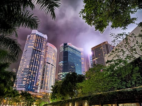 Hong Kong, China - April 06, 2024 - Tall skyscrapers in Hong Kong shine brightly against a moody sky, surrounded by lush greenery creating a vibrant urban atmosphere.