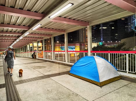 Hong Kong, China - April 06, 2024 - A tent occupied by a homeless individual is placed in a public walkway in Hong Kong, while a person walks by with a dog at night.