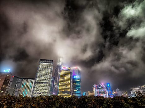 Hong Kong, China - April 06, 2024 - Tall modern skyscrapers illuminate the Hong Kong skyline at night, with clouds drifting overhead and city lights shining bright.