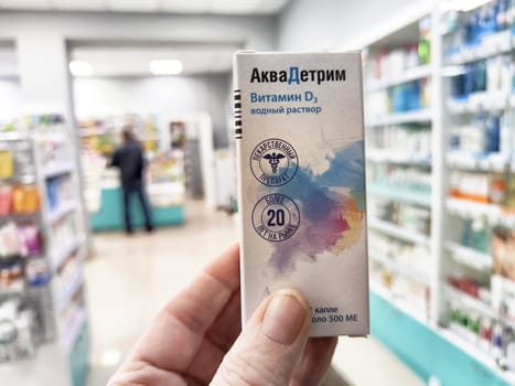 Kirov, Russia - January 10, 2025: A hand holds a vitamin D3 solution in a pharmacy, while a customer browses in the background among shelves filled with health products.