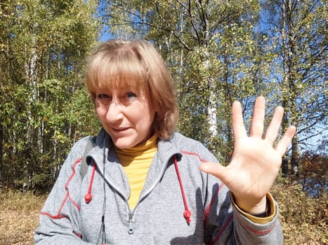 A middle-aged woman smiles and waves while exploring a beautiful autumn landscape, embracing the joy of nature.