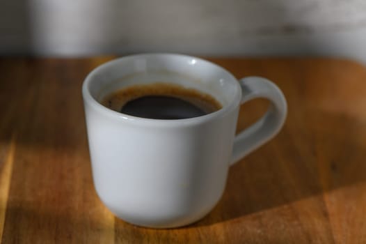 Warm sunlight bathes a small cup of dark coffee resting on a polished wooden surface, inviting a moment of relaxation.