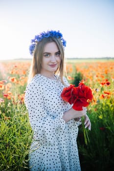 Beautiful blonde woman wearing a blue cornflower wreath poses in a poppy field holding a bouquet of red poppies