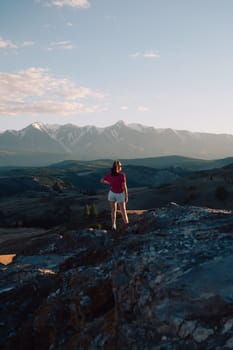 A woman in casual clothing stands on a rocky peak, gazing at scenic mountains and a vast valley under golden sunlight.
