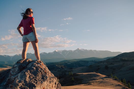A woman in casual clothing stands on a rocky peak, gazing at scenic mountains and a vast valley under golden sunlight.