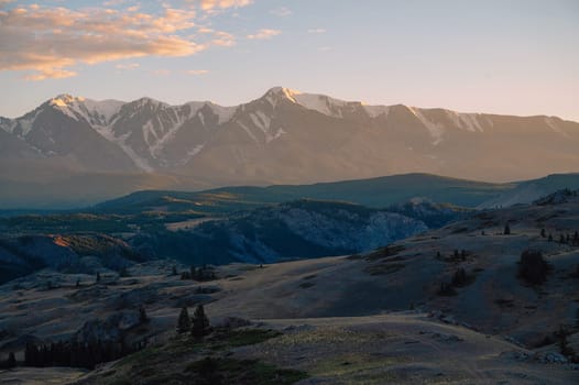 A breathtaking sunset over a rugged mountain landscape, casting warm golden light across hills, trees, and rocky terrain. Altai Mountains