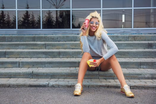 Young woman sitting on stone steps, holding a soda can and snack, enjoying a sunny day in a park setting.