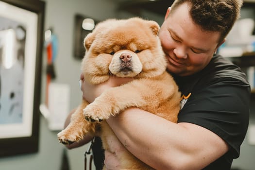 A man gently holds a fluffy dog during a grooming session in a well-lit space.