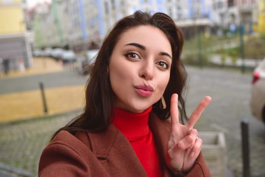 Charming smiling lady in a brown autumn coat takes a selfie on the street with a peace sign
