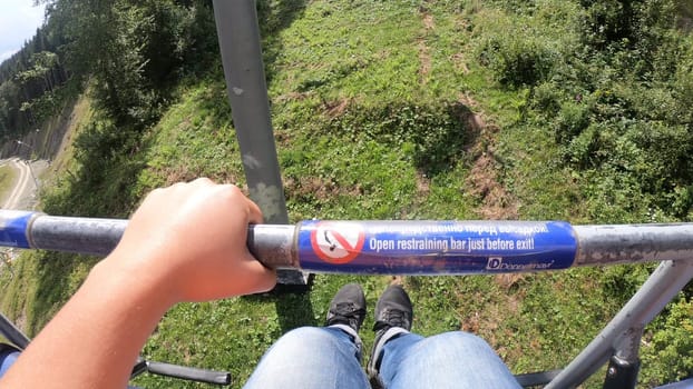 Bukovel, Ukraine 23 August 2020:-Girl rides or climbs on lift or cable car up mountain. Girl's legs hang down, holds on handle, movement of cable car seat over slope overgrown green grass close-up.