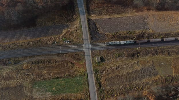 Top View Freight Train Passing Railway Crossing. Aerial Drone View Flight Over Intersection of Railway Tracks and Asphalt Road. Transportation, Rail freight, Delivery. Railway Wagon Rides on Railroad.