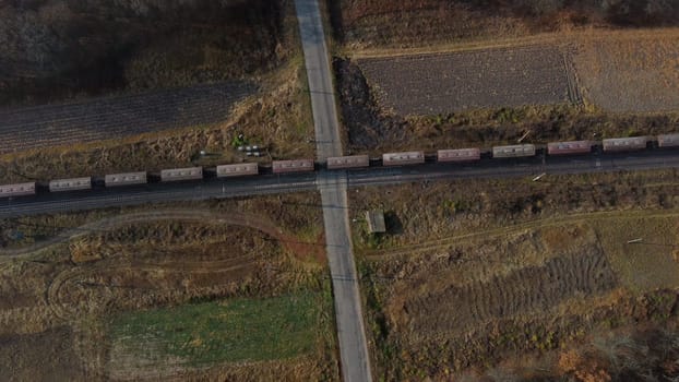 Top View Freight Train Passing Railway Crossing. Aerial Drone View Flight Over Intersection of Railway Tracks and Asphalt Road. Transportation, Rail freight, Delivery. Railway Wagon Rides on Railroad.