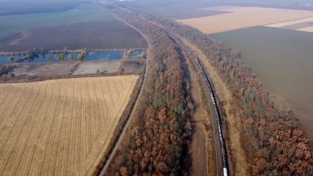 Panoramic View of Moving Freight Train Along Railway Tracks Among Trees Between Agricultural Fields on an Autumn Day. Landscape Freight Cars or Railway Wagon Rides on Railroad. Top View Rail freight