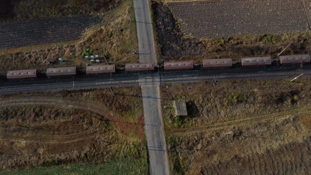 Top View Freight Train Passing Railway Crossing. Aerial Drone View Flight Over Intersection of Railway Tracks and Asphalt Road. Transportation, Rail freight, Delivery. Railway Wagon Rides on Railroad.