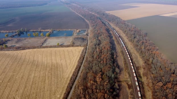 Panoramic View of Moving Freight Train Along Railway Tracks Among Trees Between Agricultural Fields on an Autumn Day. Landscape Freight Cars or Railway Wagon Rides on Railroad. Top View Rail freight