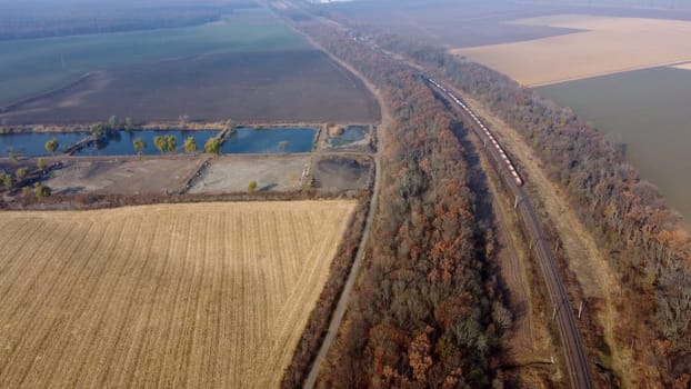 Panoramic View of Moving Freight Train Along Railway Tracks Among Trees Between Agricultural Fields on an Autumn Day. Landscape Freight Cars or Railway Wagon Rides on Railroad. Top View Rail freight