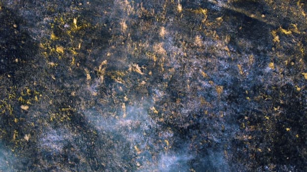 Aerial drone view flight over black ash of field of burned brown yellow dry grass and white smoke on sunny autumn day. View from above. Texture natural background of ecological environmental disaster.