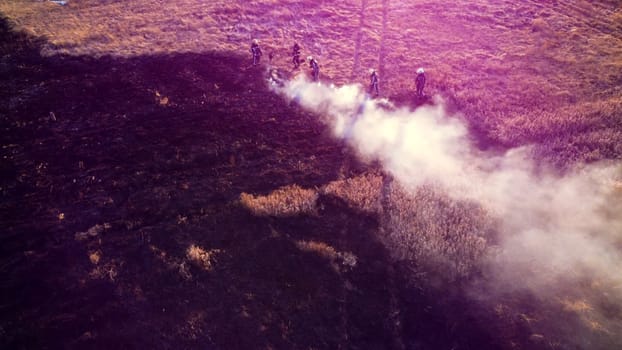 Group of firefighters puts out flame fire due to burning of dry grass in field on sunny day. View from above. Aerial drone view. Old dry grass burning on field. White smoke rising. Red pink sun glare