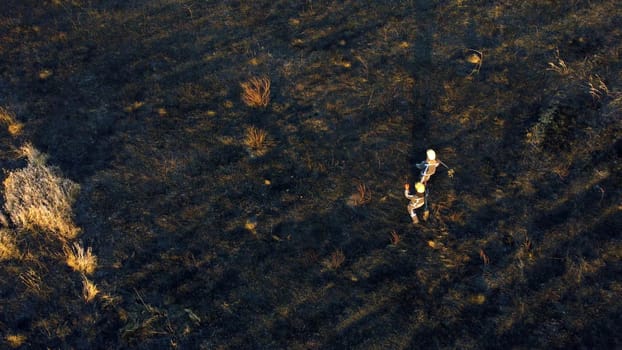Two firefighters walk on black scorched earth after fire and burning dry grass in field. Two people in uniforms of firefighters walk on black earth and ash after extinguishing fire. Aerial drone view