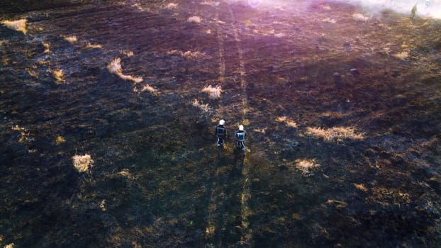 Two firefighters walk across a scorched field of black ash. View from above. Aerial drone view. Firemen walking on the burnt black grass, through conflagration on sunny summer day. Red pink sun glare