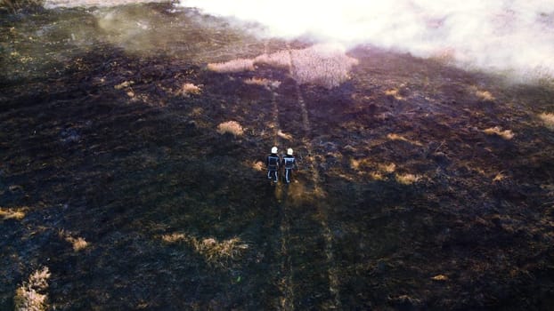 Two firefighters walk on black scorched earth after fire and burning dry grass in field. Firefighters walking on black land, ash after extinguishing fire. Aerial drone view. Red sun glare. Sun beams