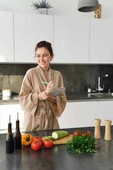 Vertical shot of brunette woman in bathrobe, cooking in the kitchen, writing down recipe in notebook, standing near chopping board with vegetables, making salad and smiling.