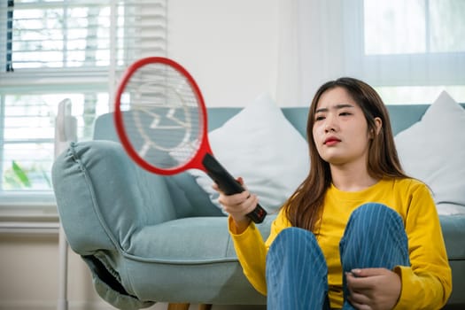 Asian female sitting floor using mosquito device swatter or electric net racket, Young woman killing mosquitoes hand holding fly swatter like weapon in living room at home, technology insect killer