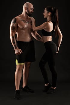 Athletic bald, tattooed fellow in black shorts and sneakers with beautiful brunette lady in leggings and top are posing standing sideways on black background and looking at each other, hugging. Fitness couple, chic muscular bodies, gym concept. The love story.