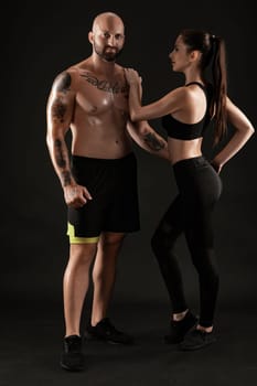 Athletic bald, tattooed man in black shorts and sneakers with beautiful brunette maiden in leggings and top are posing on black background and looking at the camera, hugging. Fitness couple, chic muscular bodies, gym concept. The love story.