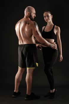 Strong bald, tattooed male in black shorts and sneakers, standing back, with charming brunette female in leggings and top are hugging on black background and looking at the camera. Fitness couple, chic muscular bodies, gym concept. The love story.