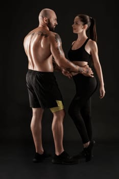 Strong bald, tattooed guy in black shorts and sneakers, standing back, with charming brunette girl in leggings and top are hugging standing sideways on black background and looking at each other. Fitness couple, chic muscular bodies, gym concept. The love story.