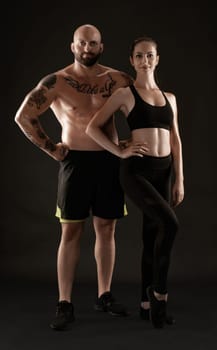 Strong bald, tattooed fellow in black shorts and sneakers with charming brunette lady in leggings and top are posing on black background and looking at the camera. Fitness couple, chic muscular bodies, gym concept. The love story.