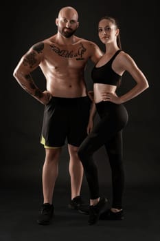 Strong bald, tattooed man in black shorts and sneakers with charming brunette maiden in leggings and top are posing on black background and looking at the camera. Fitness couple, chic muscular bodies, gym concept. The love story.