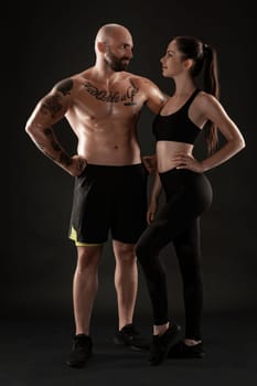 Handsome bald, tattooed man in black shorts and sneakers with gorgeous brunette woman in leggings and top are posing on black background and looking at each other. Fitness couple, chic muscular bodies, gym concept. The love story.