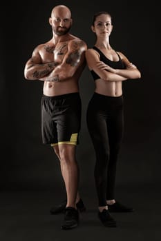 Handsome bald, tattooed guy in black shorts and sneakers with gorgeous brunette girl in leggings and top are posing with crossed hands on black background and looking at the camera. Fitness couple, chic muscular bodies, gym concept. The love story.