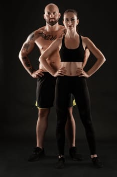 Handsome bald, tattooed fellow in black shorts and sneakers with gorgeous brunette lady in leggings and top are posing on black background and looking at the camera. Fitness couple, chic muscular bodies, gym concept. The love story.