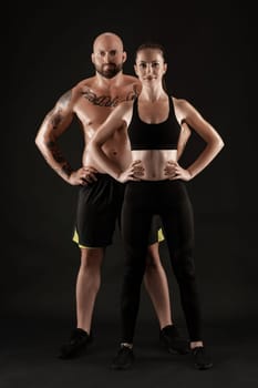 Handsome bald, tattooed man in black shorts and sneakers with gorgeous brunette maiden in leggings and top are posing on black background and looking at the camera. Fitness couple, chic muscular bodies, gym concept. The love story.