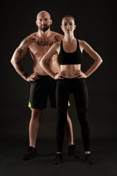 Good-looking bald, tattooed man in black shorts and sneakers with attractive brunette woman in leggings and top are posing on black background and looking at the camera. Fitness couple, chic muscular bodies, gym concept. The love story.