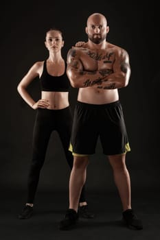 Good-looking bald, tattooed male in black shorts and sneakers with attractive brunette female in leggings and top are posing on black background and looking at the camera. Fitness couple, chic muscular bodies, gym concept. The love story.