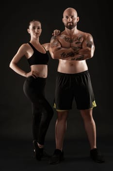 Good-looking bald, tattooed fellow in black shorts and sneakers with attractive brunette lady in leggings and top are posing on black background and looking at the camera. Fitness couple, chic muscular bodies, gym concept. The love story.