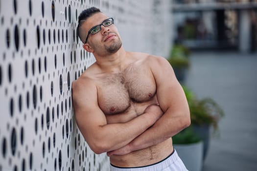 Muscular sportsman wearing glasses, crossing arms while resting against sleek metal wall after intense outdoor training session in russian urban setting