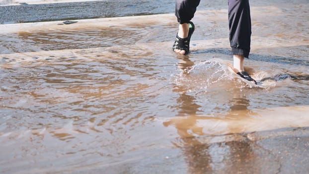 Woman wearing sandals is splashing water while walking through puddle on pedestrian crossing after rain