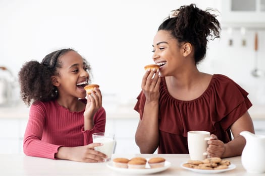 Pretty black mother and teen daughter eating cookies, happy african american girl having breakfast with her loving mom, kitchen interior. Healthy diet, calcium, vitamins in kids food concept