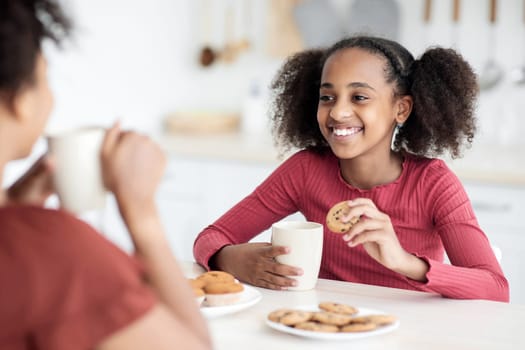 Cute african american teen girl drinking tea with her mom, sitting at kitchen table, eating biscuits, cookies, muffins, having pleasure conversation and smiling, selective focus on black kid