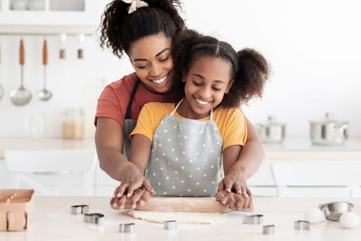 Happy black mom showing her teen kid hot to bake cookies, cheerful african american mother and daughter rolling dough for homemade pastry and smiling, wearing aprons, kitchen interior